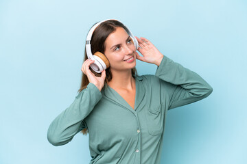 Young beautiful woman isolated on blue background in pajamas and holding a pillow and listening music
