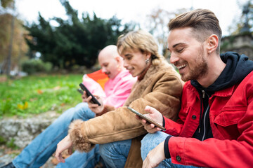 Portrait of happy young friends looking at smart phone while sitting outdoors. Caucasian people sitting at the park while using mobile phone.