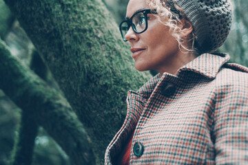 Side portrait of beautiful woman with eyeglasses wearing warm jacket and knit hat. Green trees...
