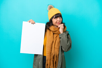 Young latin woman wearing winter jacket isolated on blue background holding an empty placard and thinking