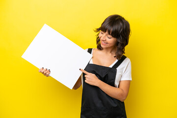 Restaurant waiter latin woman isolated on yellow bakcground holding an empty placard with happy expression and pointing it