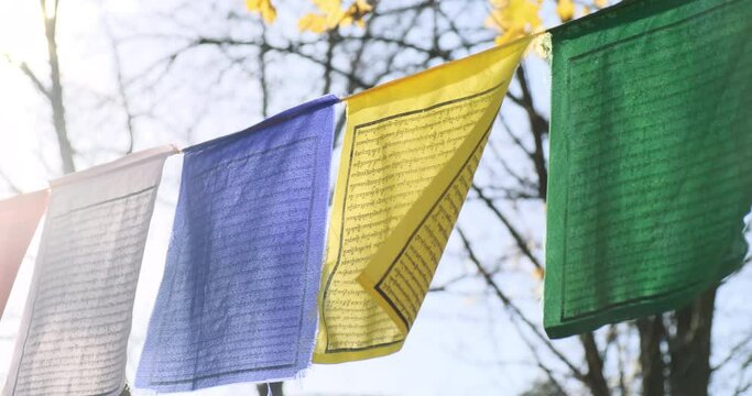 Close-up buddhist praying flags in blue sky background. Colorful buddhist tibetan prayer flag waving in autumn sun light. Buddhist pray flags slowly wind swaying in yellow trees leaves. 4k footage