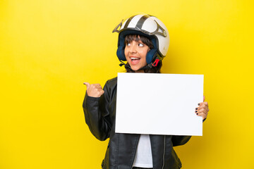 Young latin woman with a motorcycle helmet isolated on yellow background holding an empty placard and pointing side