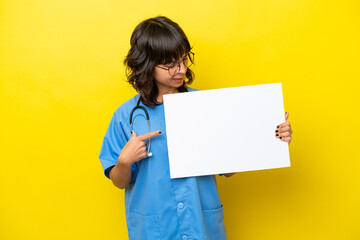 Young nurse doctor woman isolated on yellow background holding an empty placard with happy expression and pointing it