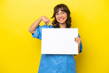 Obraz na płótnie Canvas Young nurse doctor woman isolated on yellow background holding an empty placard with happy expression and pointing it