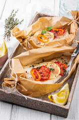 Traditional baked cod fish fillet with vegetable and herbs in backing paper served as close-up on a Nordic design plate - obrazy, fototapety, plakaty