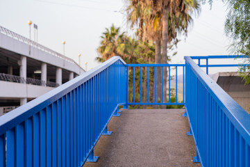 Horizontal photograph. Pedestrian bridge, no persons, white with blue railings, on asphalt highway. With clear sky, palm trees and building in the background. At sunset. Tenerife, Canary Islands Spain
