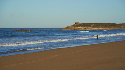 Fisherman with fishing rod on the beach. Rocks in blue sea and hills landscape. Grey sky for copy space. France, Brittany, Kerhillio.