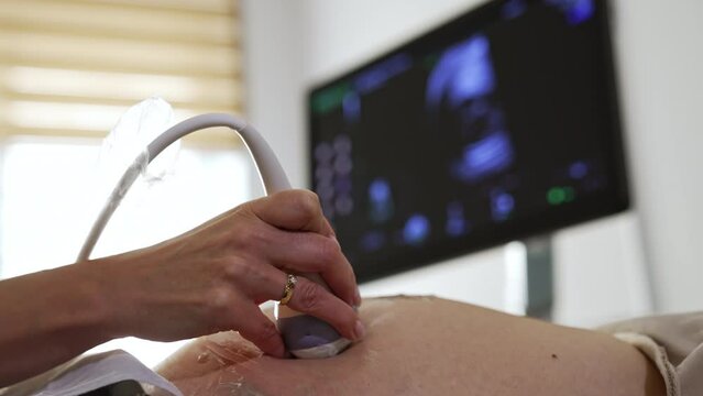 Ultrasonic diagnostic for pregnant lady. Doctor is moving the transducer by the patient’s abdomen in the hospital. Close up. Black screen at backdrop in blur.