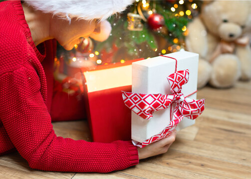 A Girl In A Red Sweater Peeks Into An Open Box With A Red Bow. Christmas Photo With A Girl In A Red Cap With A Gift From A Gift Box Pours Bright Light