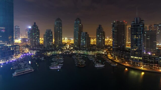 Dubai Marina during all night timelapse, Glittering lights swithcing off and tallest skyscrapers. Illuminated towers, yachts and traffic on the road on background