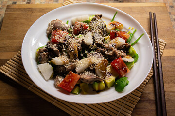 oriental salad with tuna and fresh vegetables on a white plate - standing on a makisu - with black Chinese chopsticks on a wooden cutting board background