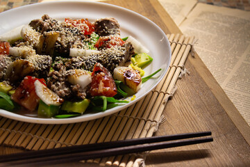 An oriental salad with tuna and fresh vegetables in a white plate stands on a bamboo makisu and black Chinese chopsticks against the background of a wooden cutting board and an old newspaper