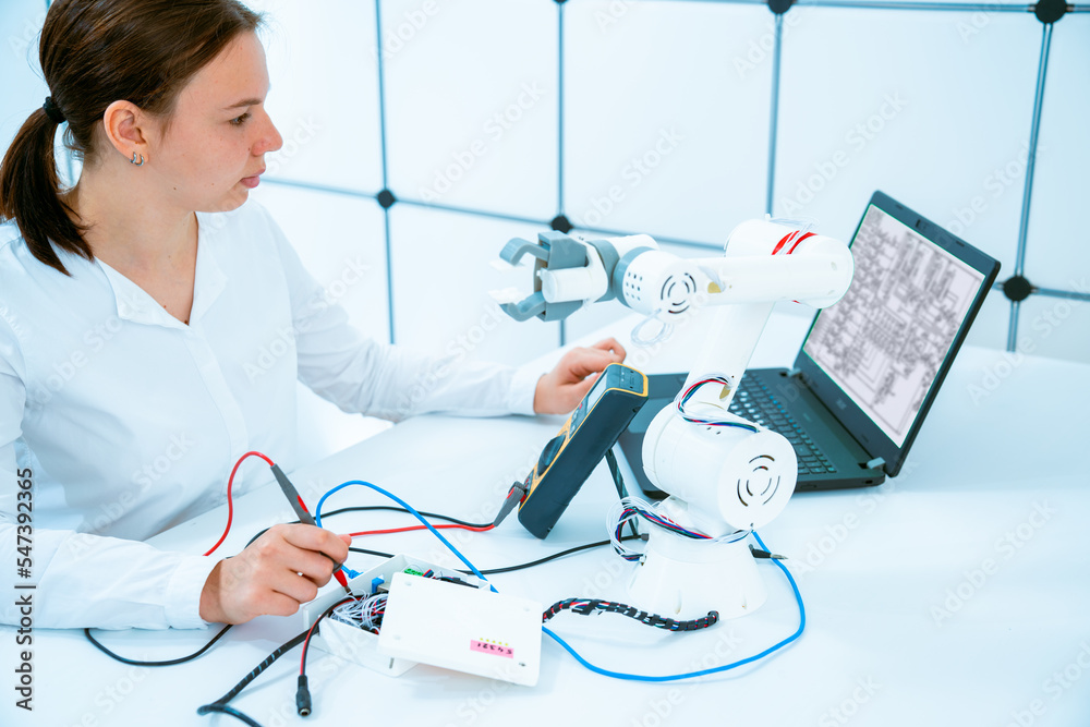 Wall mural debugging to the model of an industrial robot of the University laboratory a young woman measures the voltage in the control circuits of the robot with a tester