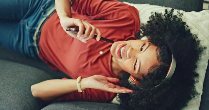 Top view, woman and on sofa singing with headphones, phone and happy in living room. African American girl, digital device and fun being relax, sing and enjoy break on couch in lounge and dancing.