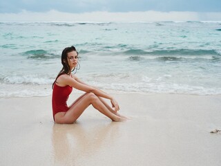 Fototapeta na wymiar Woman with a beautiful tan tourist in a red swimsuit sitting on the sand on the beach in the ocean in waves, travel and beach vacation