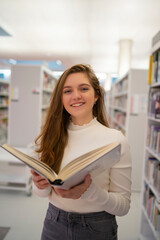 Portrait of happy beautiful young student. She is reading in modern university library.