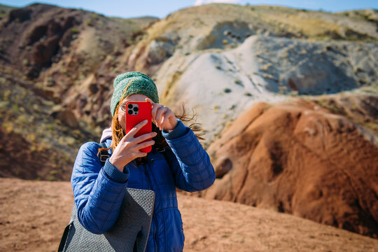 Woman traveller holding smartphone covering her face on blurred mountain background. Happy female hipster, blogger in vacation. Travel photo concept.