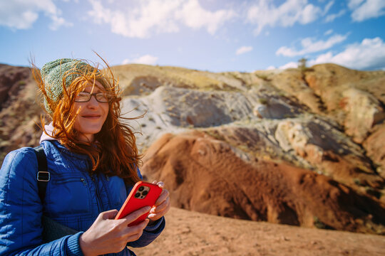 Beautiful female traveler using smartphone standing on amazing mountain background. Young long hair woman tourist enjoying activity vacation.