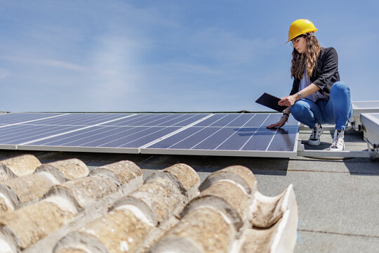 Ingegnere donna con caschetto protettivo giallo verifica le condizioni di un impianto fotovoltaico sul tetto di una casa con un tablet in mano