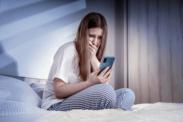 Woman crying with smartphone in bed at home or hotel in the darkness. Lady relaxing in a nightgown....