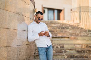 Obraz premium young handsome man with a mobile phone in his hands, a man in a white shirt holds a phone on the street in the city, a businessman with a phone