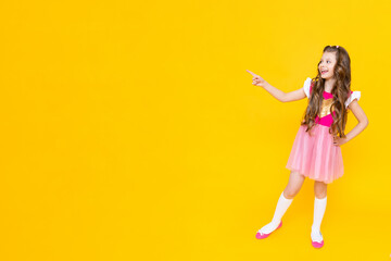 A little girl points to your ad on a yellow isolated background. A beautiful child in a pink dress points to an empty place and smiles broadly.