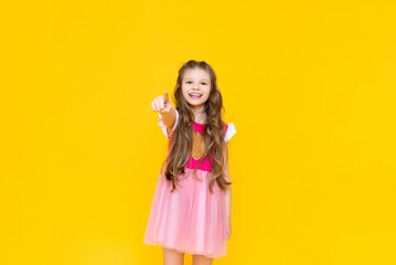 A little princess in a pink dress with curly hair smiles broadly and points forward with her index finger. A beautiful, charming child on a yellow isolated background.