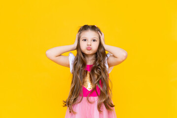 The little girl clutched her head and puffed out her cheeks against the yellow isolated background. The little princess in the pink dress is shocked.