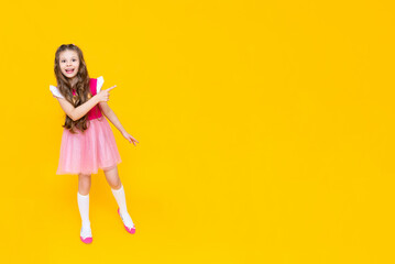 A little girl points to your ad on a yellow isolated background. A beautiful child in a pink dress points to an empty place and smiles broadly.