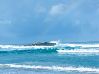 Turquoise beach with rocks and rainbow in winter with clouds and blue sky, yellow sand