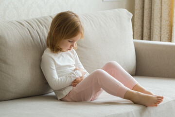 Little girl sitting on sofa and touching painful belly with arms at living room. Child suffering from stomach ache at home. - obrazy, fototapety, plakaty