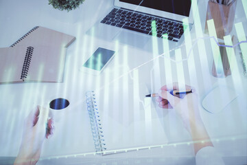 Hands writing information about stock market in notepad. Forex chart holograms in front. Concept of...