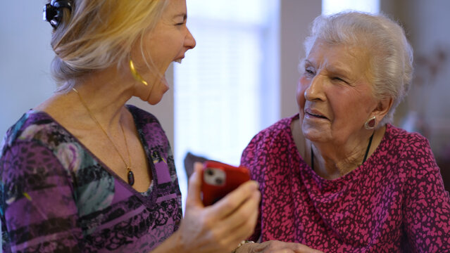 Happy laughing elderly senior woman sitting at table with middle aged daughter, watching family or journey photos on phone. Smiling blonde lady listening to older, using smartphone.