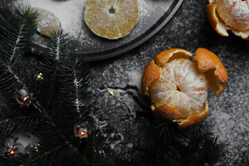 sweet orange juicy tangerines in a peeled peel lie on a black background and a black kitchen tray next to a spruce Christmas branch and sprinkled with white powder.for cards, banners, flyers, invitati