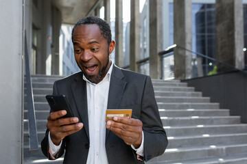 Happy african american businessman outside office building using phone and bank credit card for online shopping and money transfer, mature boss in business suit smiling.