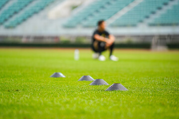 An obstacle cone set on the grass pitch for dribbling training with a player resting as blur background. Professional football training concept, close-up and selective focus on the front cone.  - Powered by Adobe