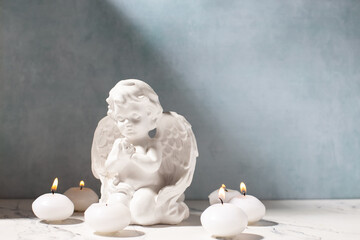 Little statue of white angle and white burning candles against blue textured wall. Selective focus.