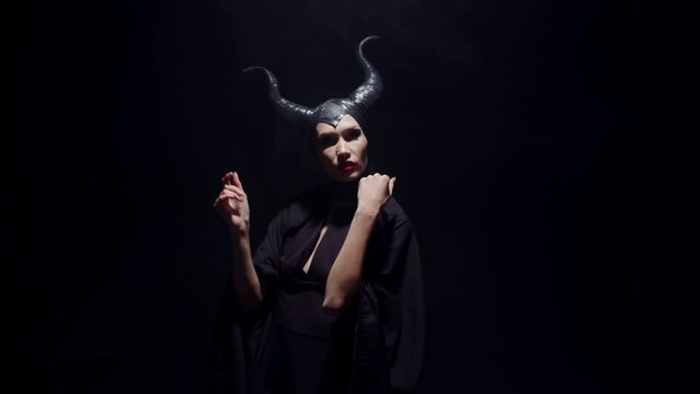 Young caucasian woman with bright makeup dressed like Maleficent slowly moving in a dark room. Beautiful woman with horns on her head showing off her mystery devil halloween outfit indoor