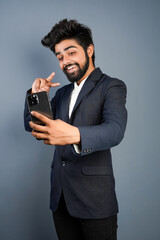Portrait of a handsome happy young businessman wearing suit standing isolated over gray background, using mobile phone, video call concept