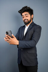Portrait of a handsome happy young businessman wearing suit standing isolated over gray background, using mobile phone, video call concept