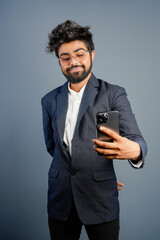 Portrait of a handsome happy young businessman wearing suit standing isolated over gray background, using mobile phone, video call concept