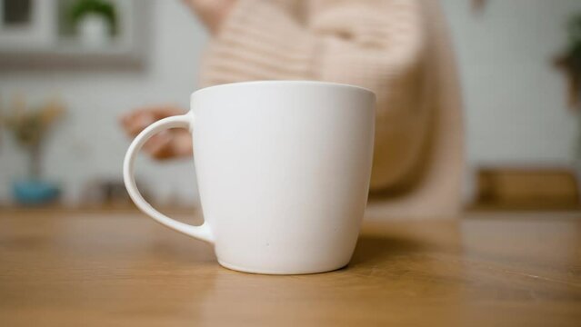Woman puts white ceramic cup on wooden table in kitchen. Cup for tea and coffee. Modern kitchen design. Housewife with manicure. Girl leaves empty cup and walks away. Dishes for liquids and hot drinks