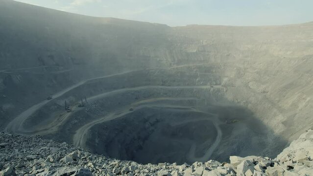 General view of a deep dusty quarry with many circular roads inside and a moving truck which seems small. Laden dumper driving across huge quarry in the far distance on sunny day