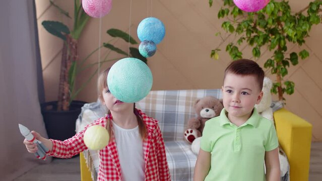 Children play astronauts in the room. A little boy touches the spheres with his hand. A little girl controls a toy rocket with her hand. The room is decorated with solar system and planets.