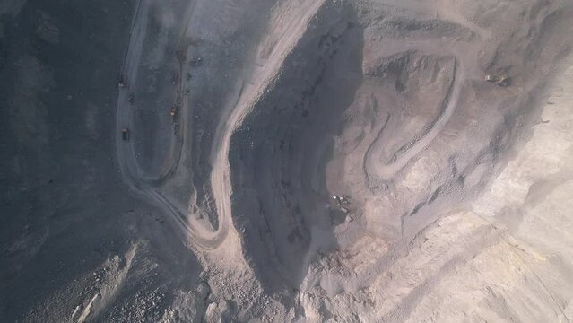 Aerial view from above of a deep dusty quarry with many narrow curved roads inside and a few moving trucks which seems small. Laden mining trucks working inside opencast mine on sunny day