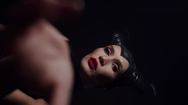 Young caucasian woman close up with bright makeup dressed like Maleficent slowly stretching forward her hands in a dark room on vertical video. Handsome woman with horns on her head showing off her