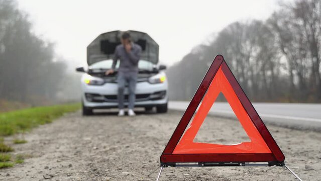 Road side warning triangle, warning oncoming traffic of a broken down car, with a man using his cell phone to call for assistance. A man near a broken car on a foggy road