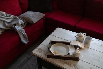 messy dark living room home with red couch, gray blanket, white farmhouse table, tea pot, mug, and dirty plates