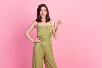 Photo of girlish adorable gorgeous woman wear khaki overall impressed pointing empty space hand on waist isolated on pink color background